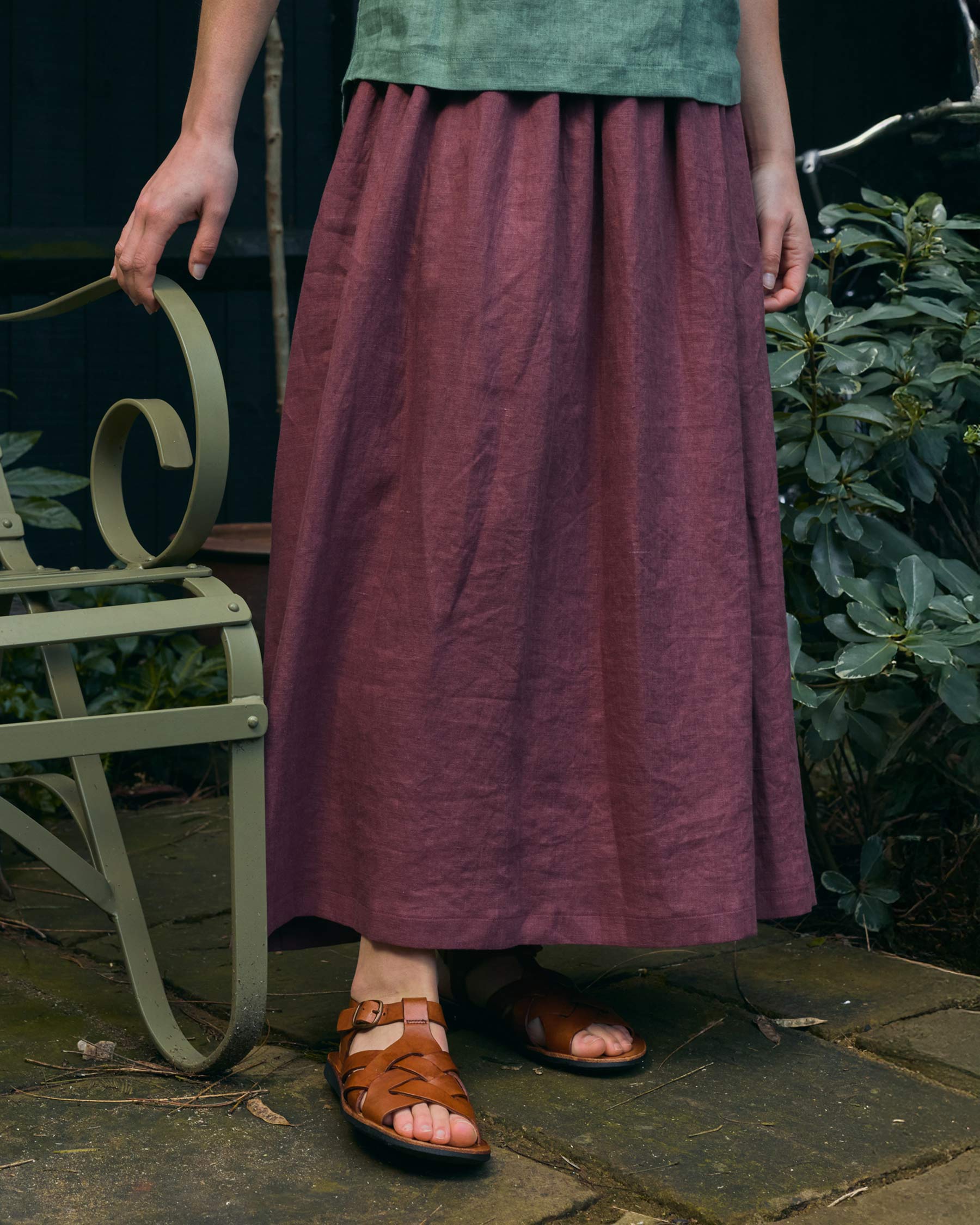 Someone wearing the Linen Maxi Length Skirt No.12 in Wild Ginger from Pyne and Smith Clothiers, paired with a green top and brown sandals, stands by a green metal chair on a stone patio with leafy plants behind. Their face is not shown.