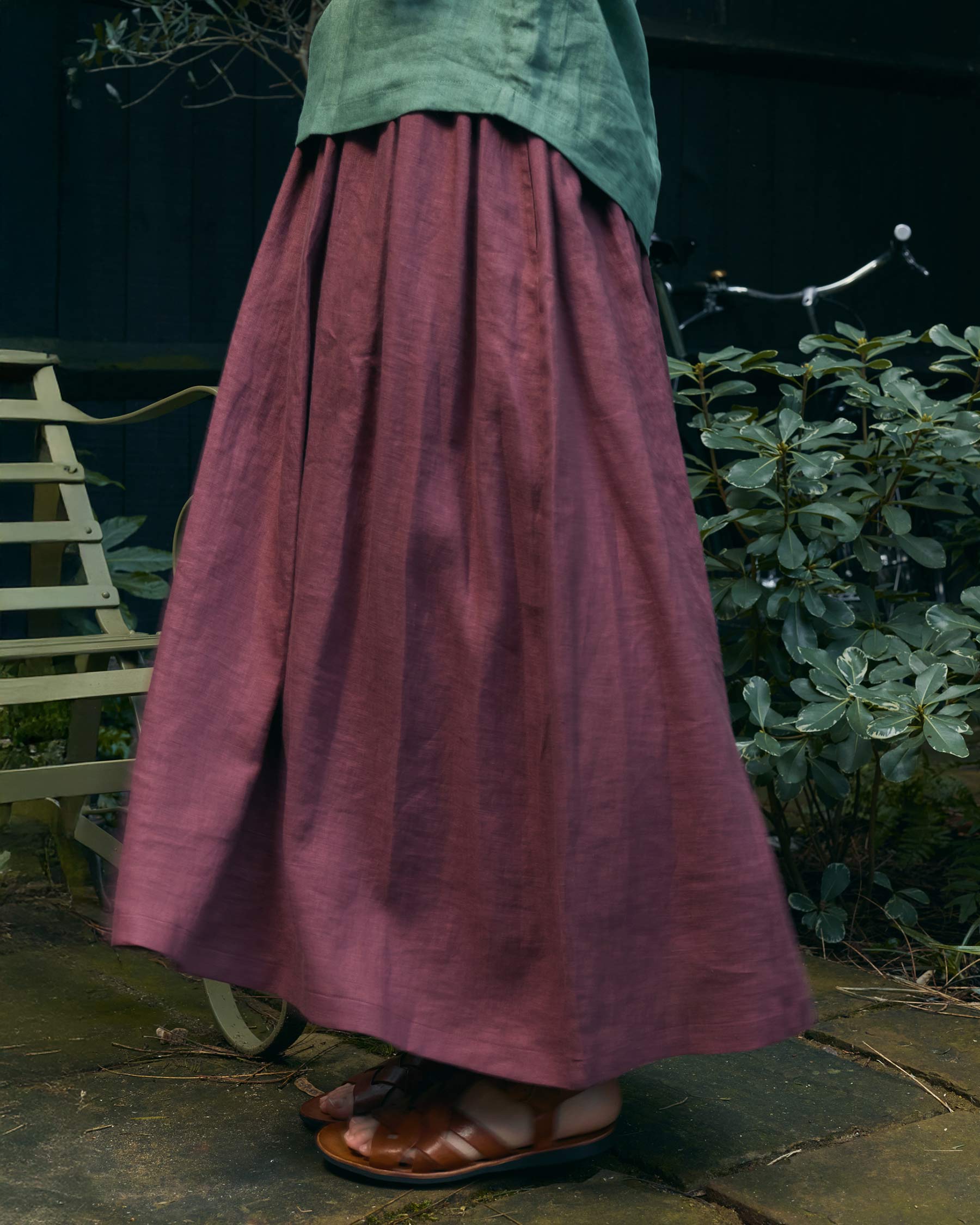 A person wearing the Pyne and Smith Clothiers Linen Maxi Length Skirt No.12 in Wild Ginger stands outdoors near a metal chair and leafy plants. The cropped photo shows only the lower half of their body.