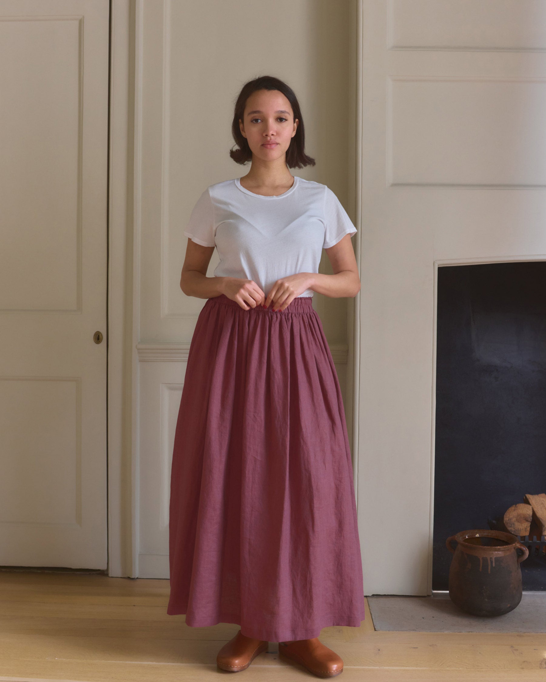 A woman stands indoors facing forward, wearing a white t-shirt and the Linen Maxi Length Skirt No.12 in Wild Ginger by Pyne and Smith Clothiers. She holds the skirts waistband in front of a cream-paneled wall and fireplace, with a neutral expression.
