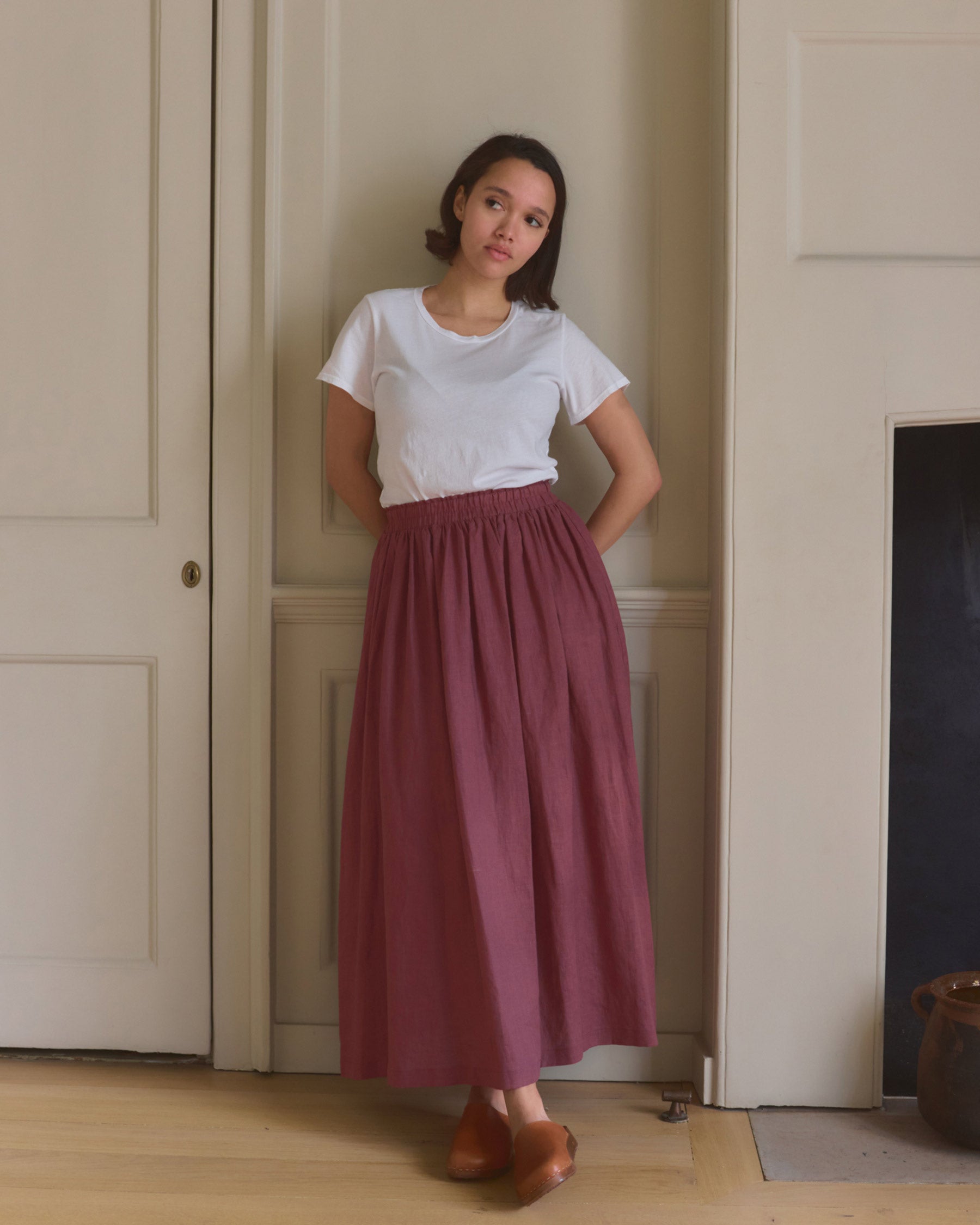 A woman stands indoors by a cream wall, wearing a white t-shirt and the Linen Maxi Length Skirt No.12 in Wild Ginger by Pyne and Smith Clothiers, paired with brown shoes. Her hands are behind her back and she has a neutral expression.