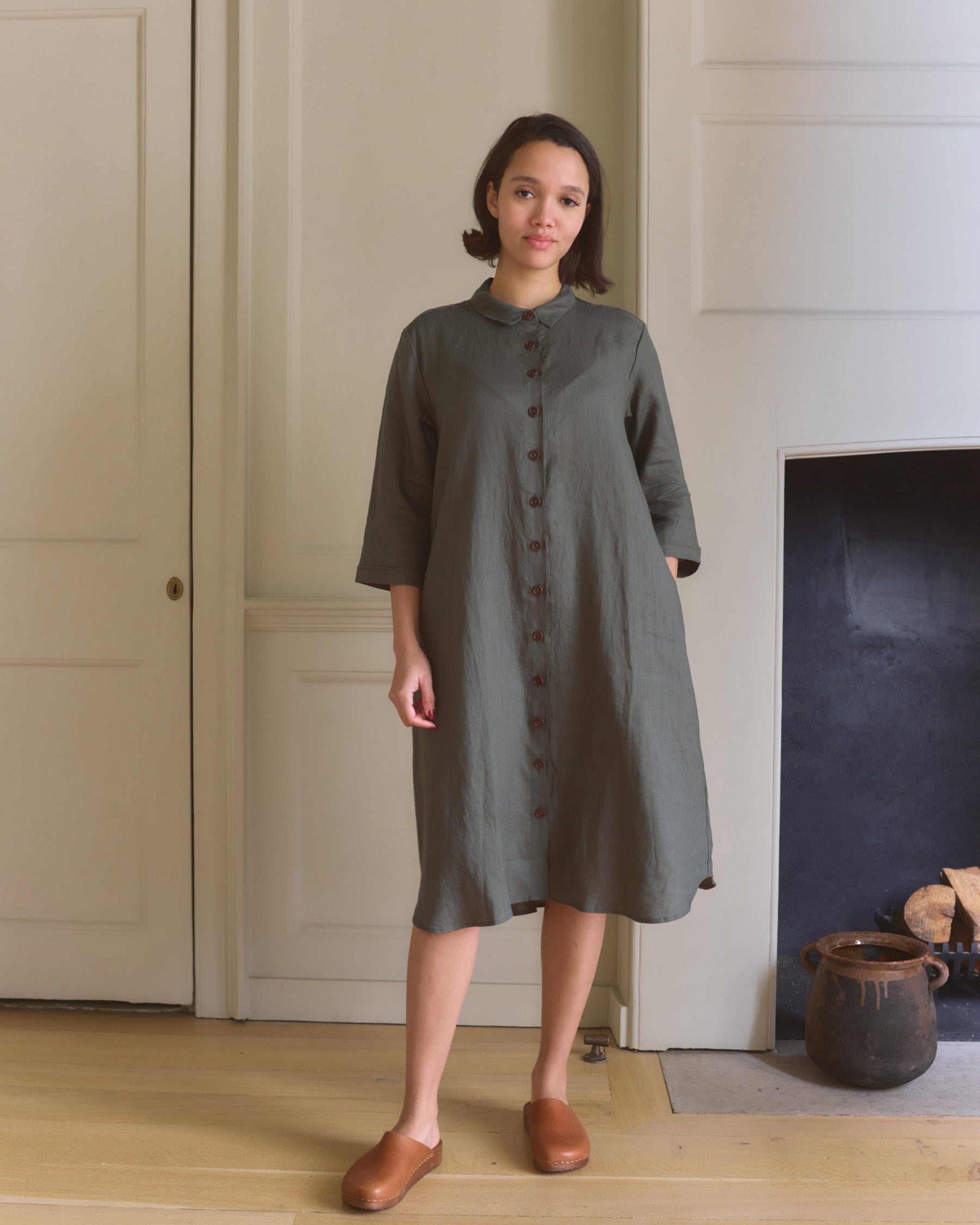 A woman stands indoors in front of pale wood paneling and a fireplace, wearing Pyne and Smith Clothiers No.39 The Linen Shirtdress in Beetle Green with brown slip-on shoes. Pottery decorates the room.
