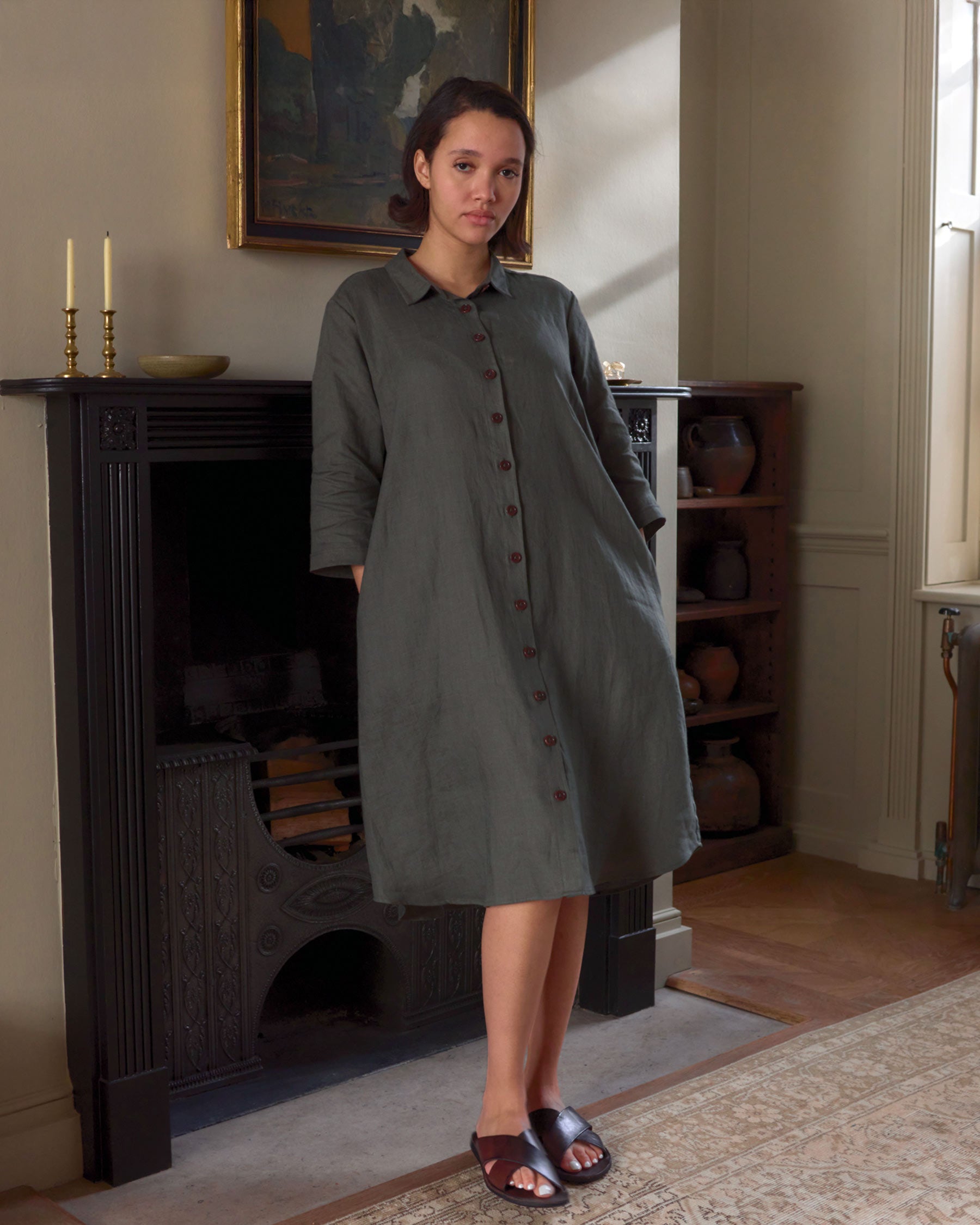 A woman wearing the No.39 The Linen Shirtdress in Beetle Green by Pyne and Smith Clothiers stands barefoot before a black fireplace in a softly lit, vintage-style room.