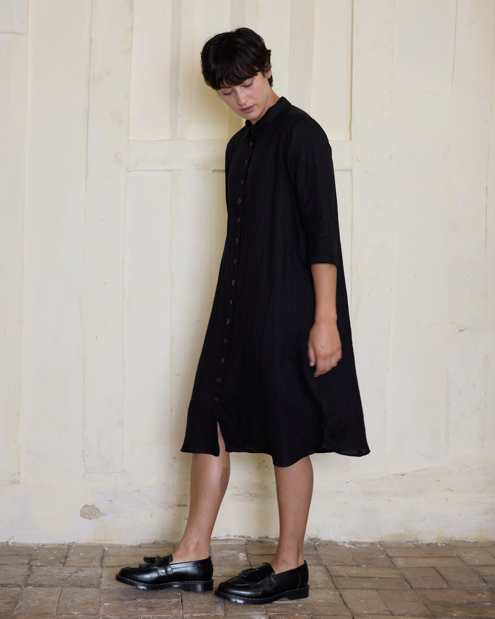Wearing the No.39 The Linen Shirtdress in Black by Pyne and Smith Clothiers, a person with short dark hair and black loafers stands indoors on a tiled floor, gazing down with a neutral expression against a pale wall.