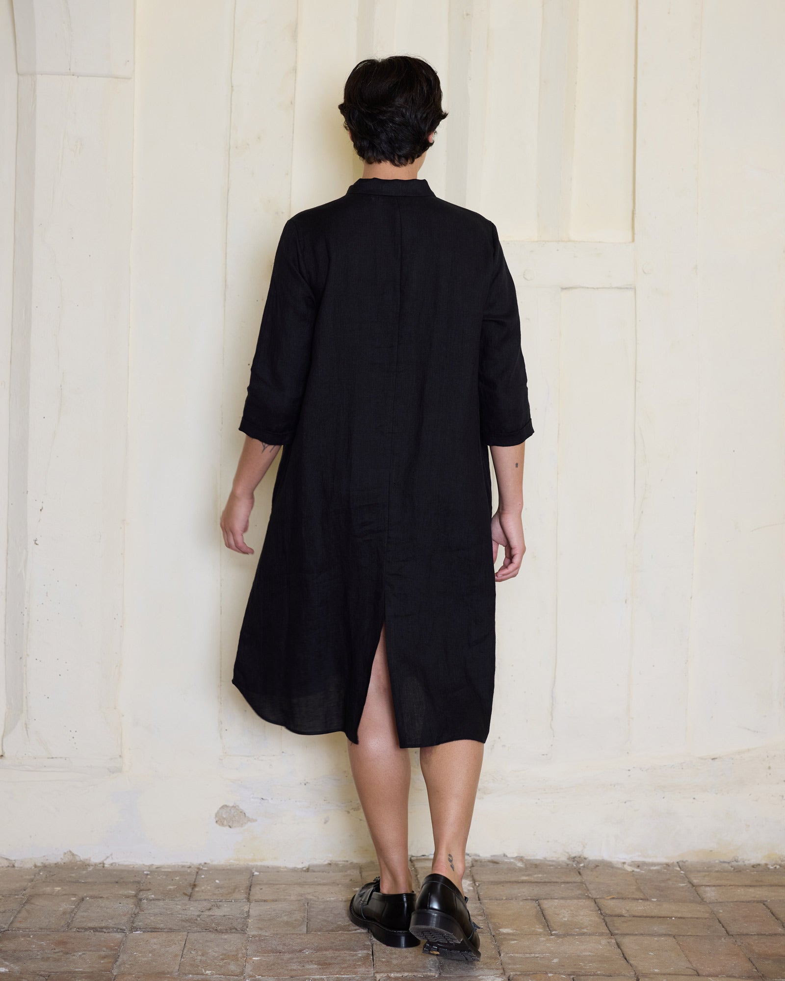 A person stands indoors against a beige paneled wall, wearing the Pyne and Smith Clothiers No.39 The Linen Shirtdress in Black, a knee-length linen shirt dress with three-quarter sleeves and a back slit, paired with black shoes on a tiled floor.
