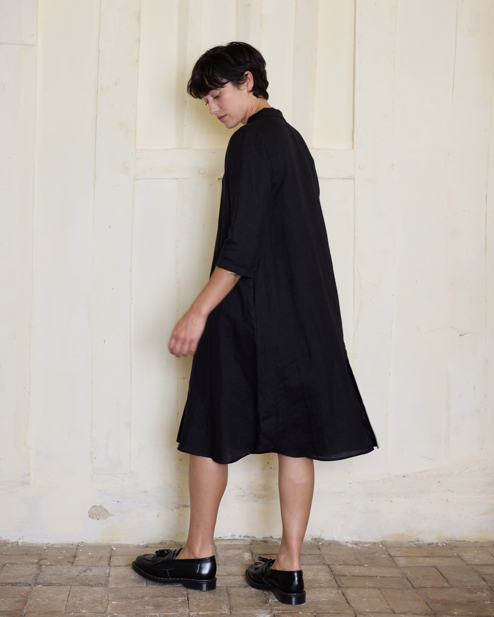 A person with short dark hair wears the No.39 The Linen Shirtdress in Black by Pyne and Smith Clothiers, paired with black shoes, standing on a tiled floor and looking over their shoulder against a light-colored textured wall.