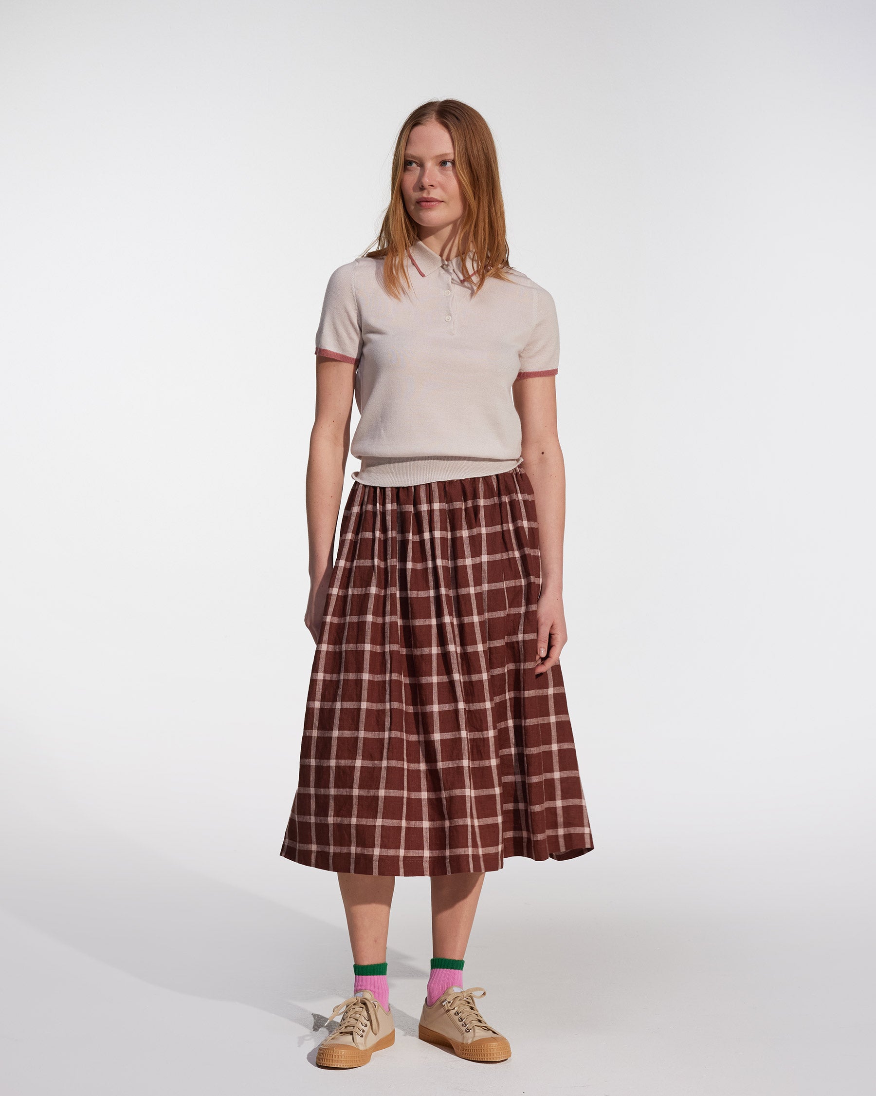 A woman stands against a white background wearing Pyne and Smith Clothiers' Linen Skirt No.11 in Neapolitan, paired with a short-sleeved light beige polo, pink socks, tan sneakers, and has straight shoulder-length hair.