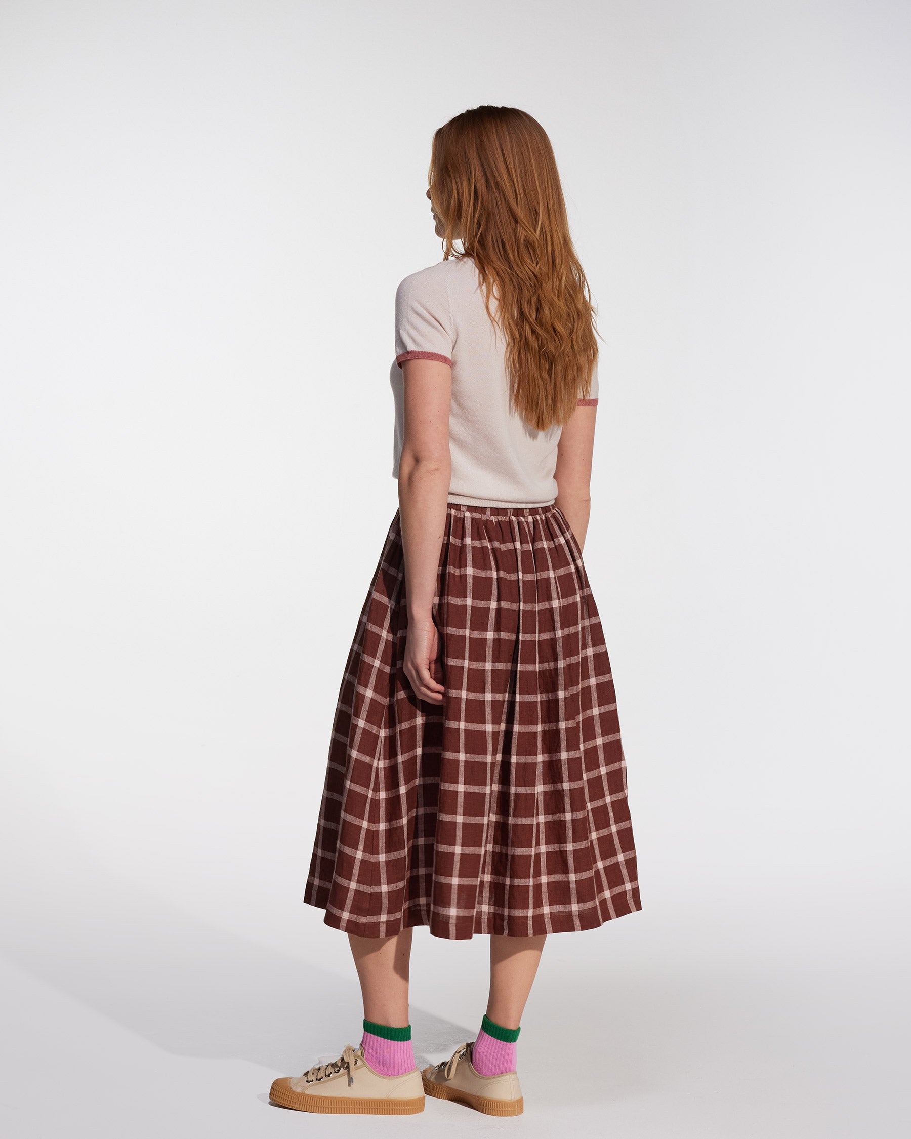 A woman with long red hair, facing away, wears a white short-sleeve top and the Pyne and Smith Clothiers Linen Skirt No.11 in Neapolitan—a brown and pink plaid linen midi skirt with an elastic waistband—plus tan sneakers and colorful socks. Plain white background.