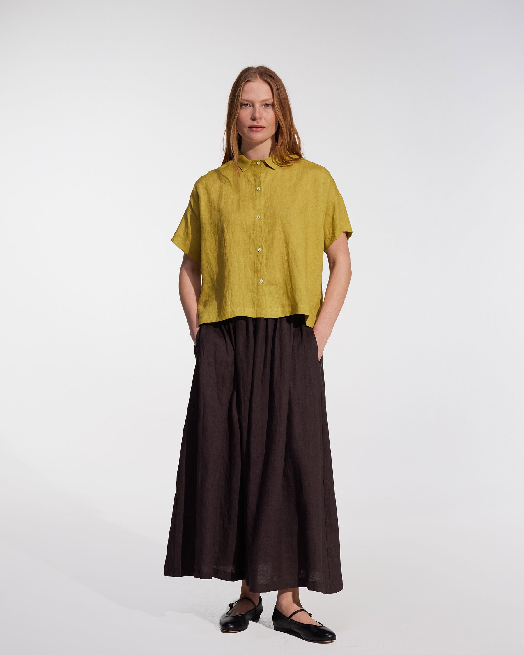 A woman stands facing forward wearing Pyne and Smith Clothiers’ Linen Maxi Skirt No.12 in Chocolate Plum with pockets, paired with a short-sleeved yellow button-up shirt, black flats, and straight light brown hair against a plain background.