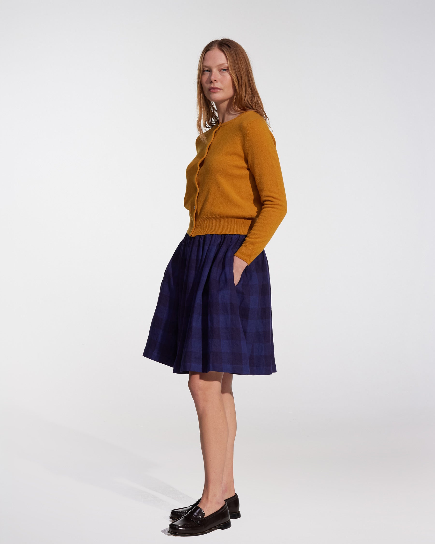 A woman stands against a white background, wearing the Pyne and Smith Clothiers Linen Skirt No.14 in Midsommar Night Check with her hands in her skirt pockets, looking slightly to the side.