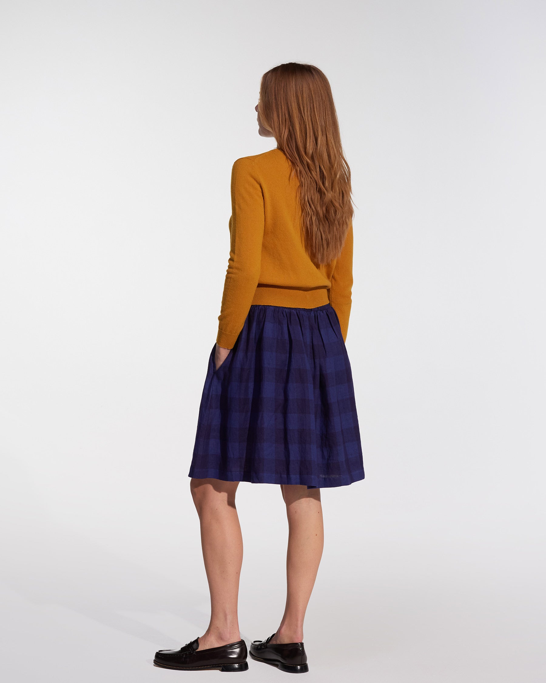 A woman with long, straight hair faces away, wearing a mustard yellow sweater, black loafers, and the Pyne and Smith Clothiers Linen Skirt No.14 in Midsommar Night Check against a plain white background.