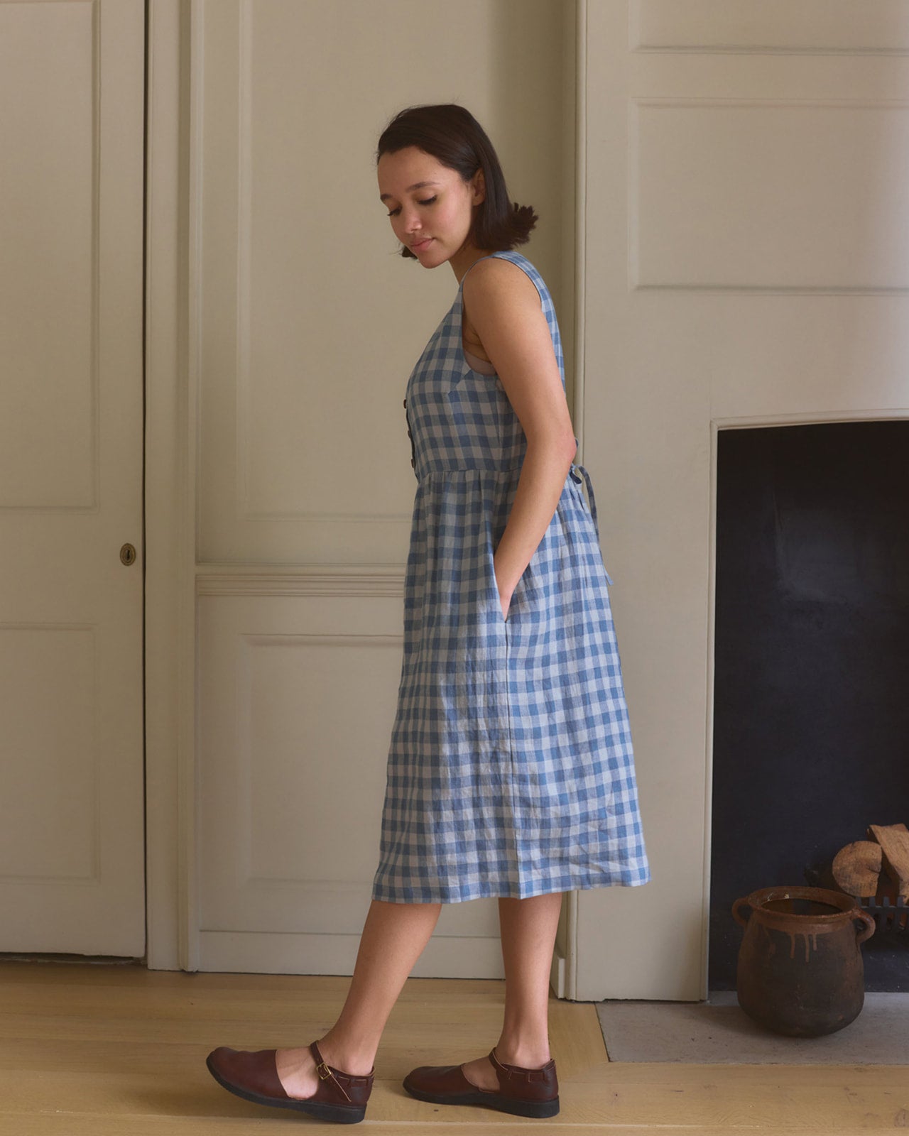 A woman in the Pyne and Smith Clothiers No.24 Sleeveless, button up Linen Dress in Bothy Check stands sideways in a light-walled room with wooden floors, hands in pockets, looking down and wearing brown flats.