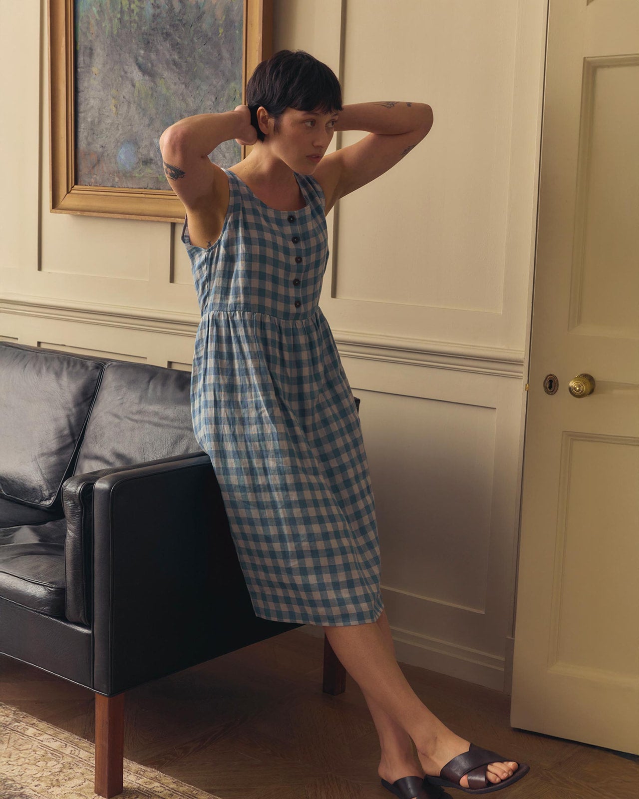 A person in the Pyne and Smith Clothiers No.24 Sleeveless, button-up Linen Dress in Bothy Check stands by a couch, arms raised to their head, in a softly lit room with a painting and beige walls.