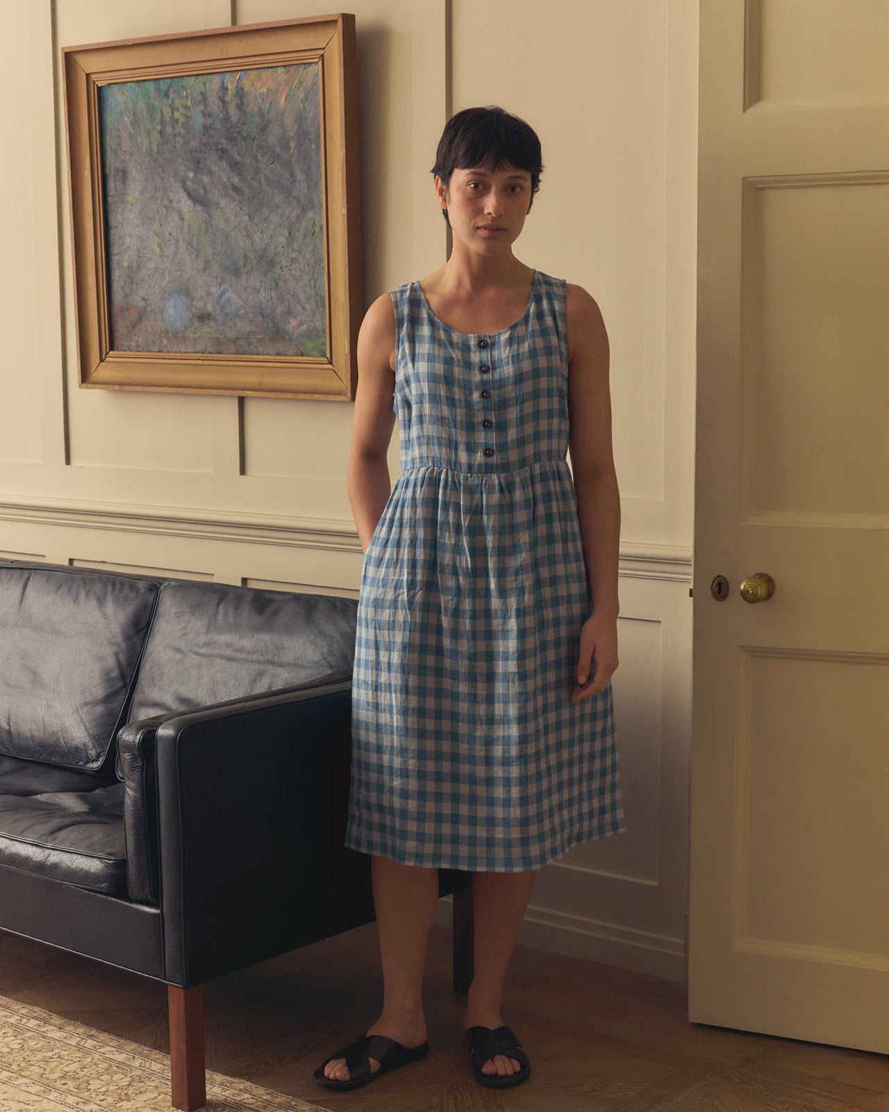 A person in the Pyne and Smith Clothiers No.24 sleeveless, button-up linen dress in Bothy Check stands indoors by a black sofa, near a beige wall with a door and framed abstract art, wearing black sandals.