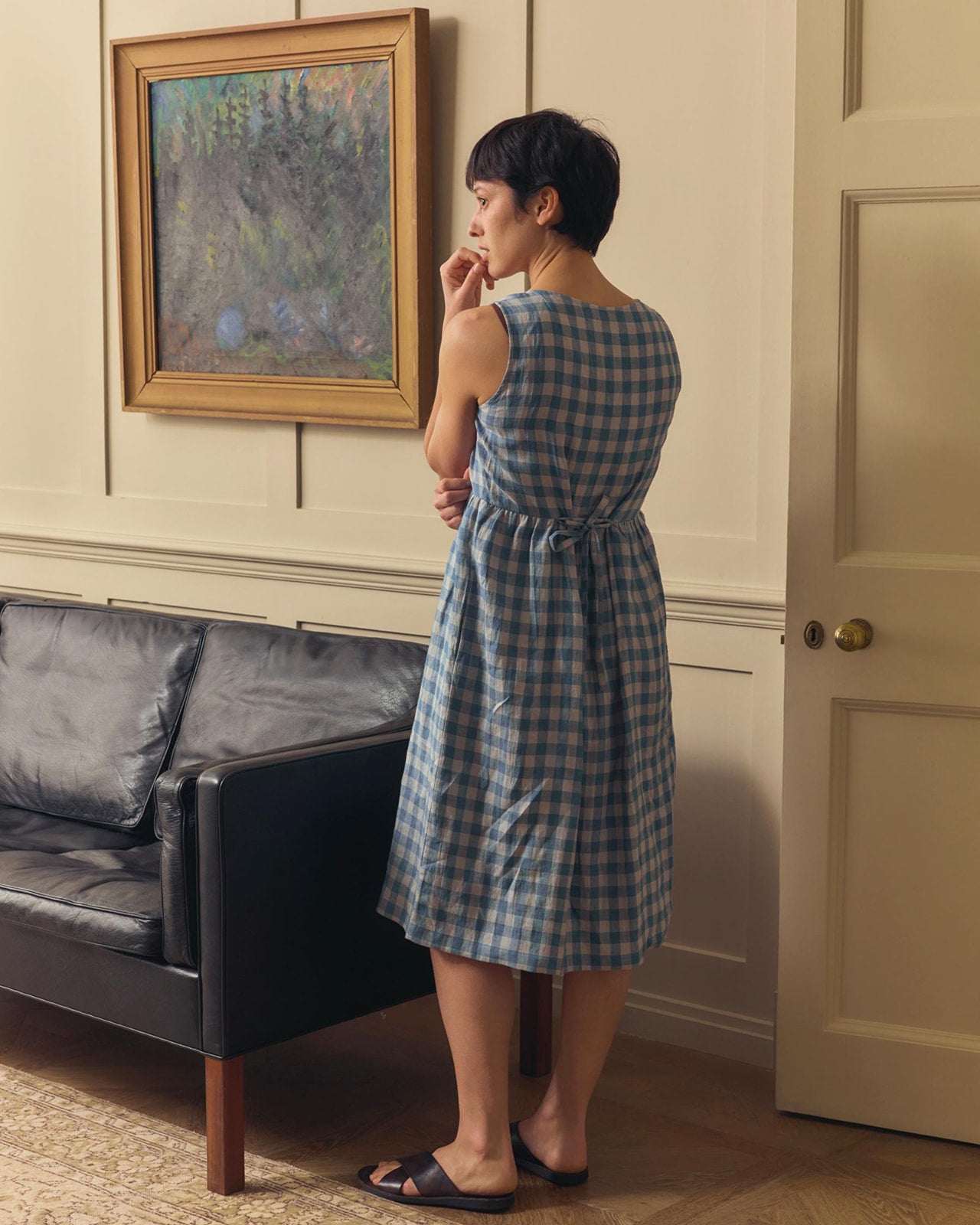 A woman wears the No.24 Sleeveless, button-up Linen Dress in Bothy Check by Pyne and Smith Clothiers, standing by a black sofa against a beige wall with framed art, looking thoughtful with one hand near her mouth.