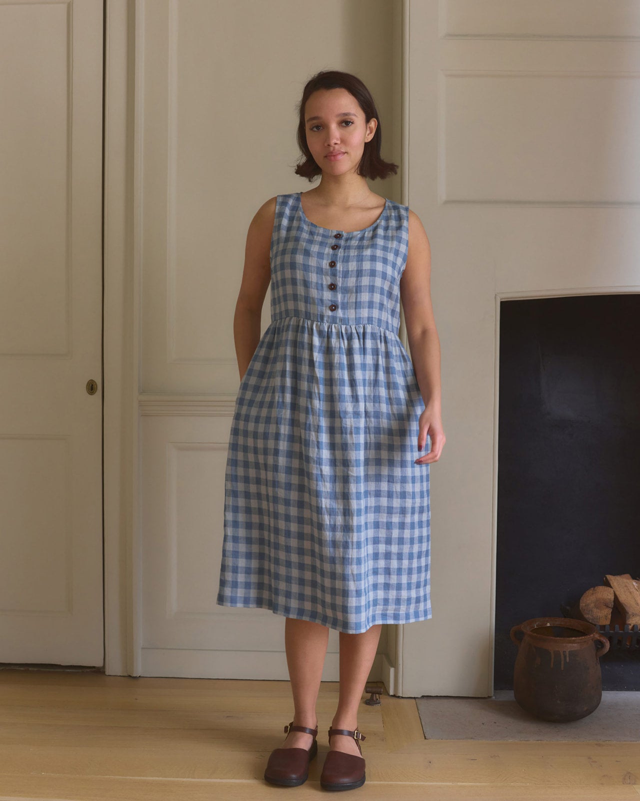 A woman stands indoors in the Pyne and Smith Clothiers No.24 sleeveless, button-up Linen Dress in Bothy Check, featuring an adjustable waist tie, paired with brown clogs by a white paneled wall and fireplace.