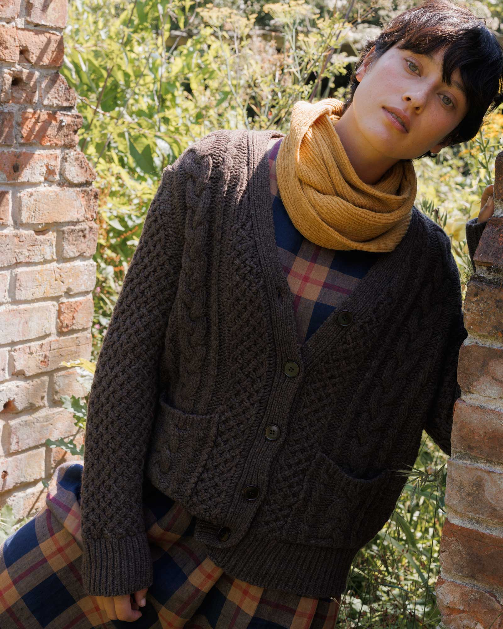 A person with short dark hair wears the Pyne and Smith Clothiers Merino Wool Cable Knit Cardigan with Pockets in Walnut, paired with a yellow scarf and plaid dress, standing outside between brick walls and greenery.
