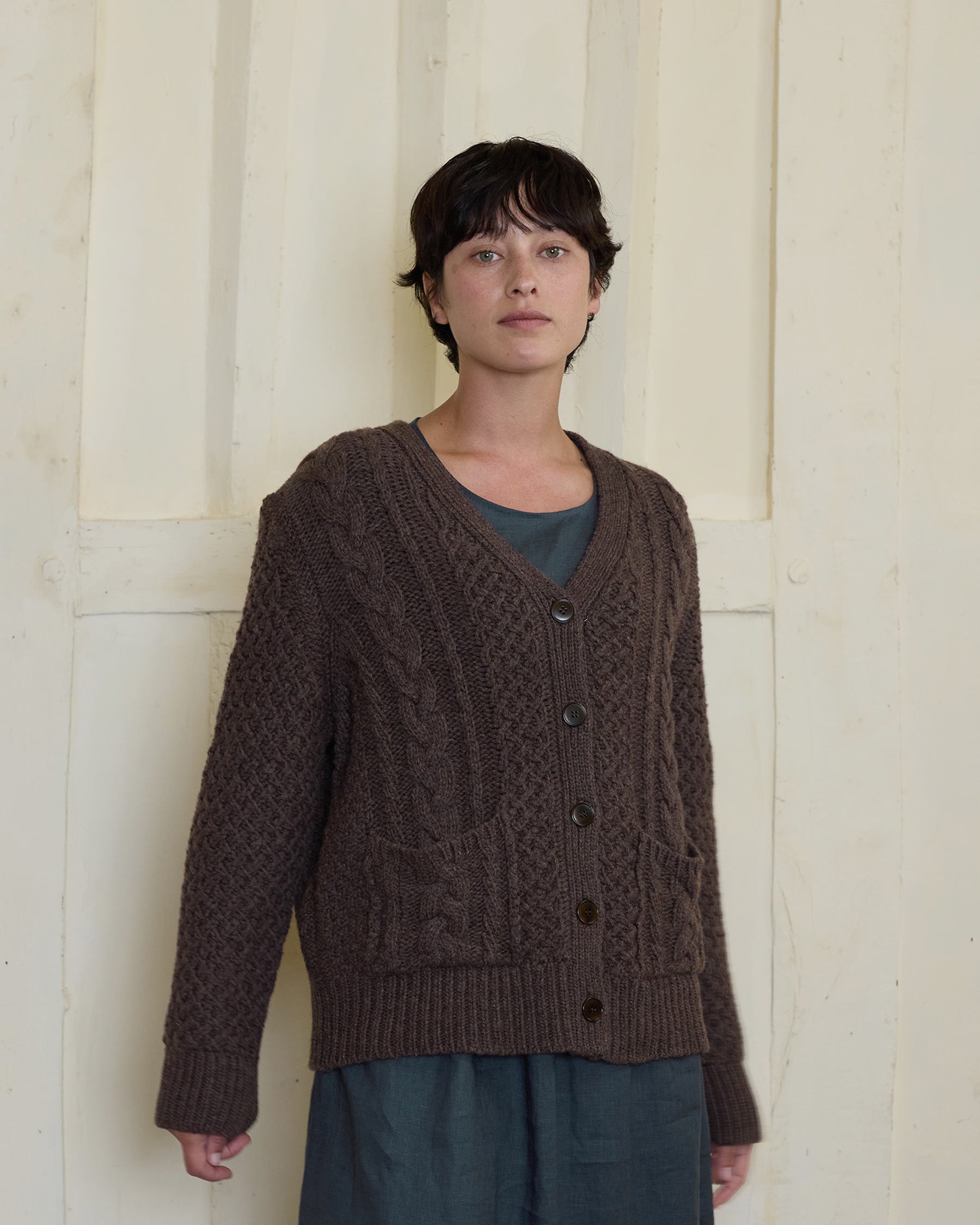 A person with short dark hair stands before a cream-paneled wall, wearing the Pyne and Smith Clothiers Merino Wool Cable Knit Cardigan with Pockets in Walnut over a gray dress.