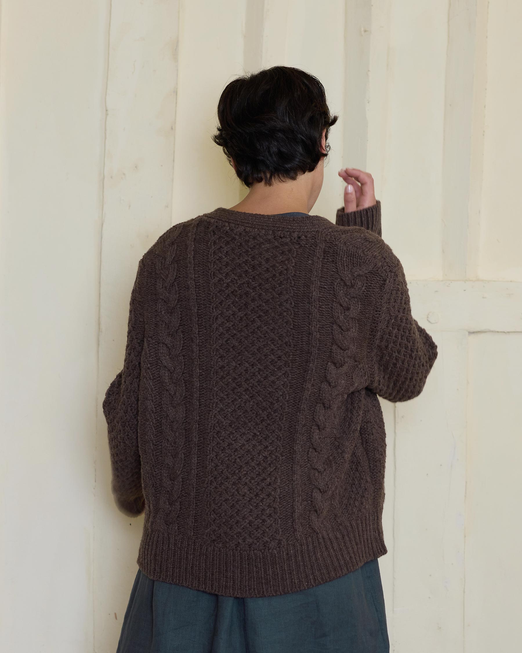 A person with short dark hair faces away, wearing the Pyne and Smith Clothiers Merino Wool Cable Knit Cardigan with Pockets in Walnut, standing against a cream-colored paneled wall.