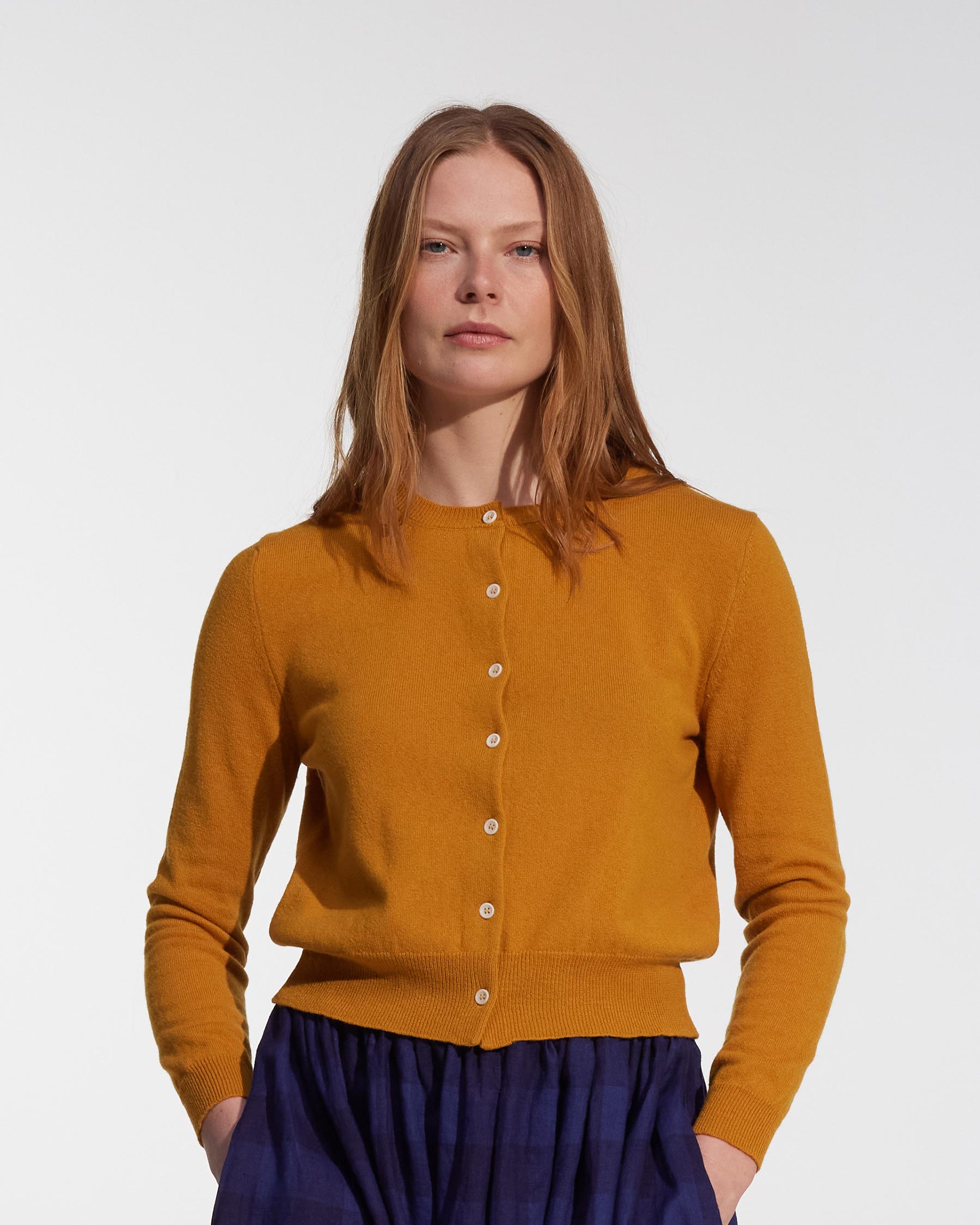 A woman with straight, light brown hair wears the Pyne and Smith Clothiers Merino Cardigan in Coleman's Yellow and a dark blue skirt, standing against a plain white background with her hands in her skirt pockets.