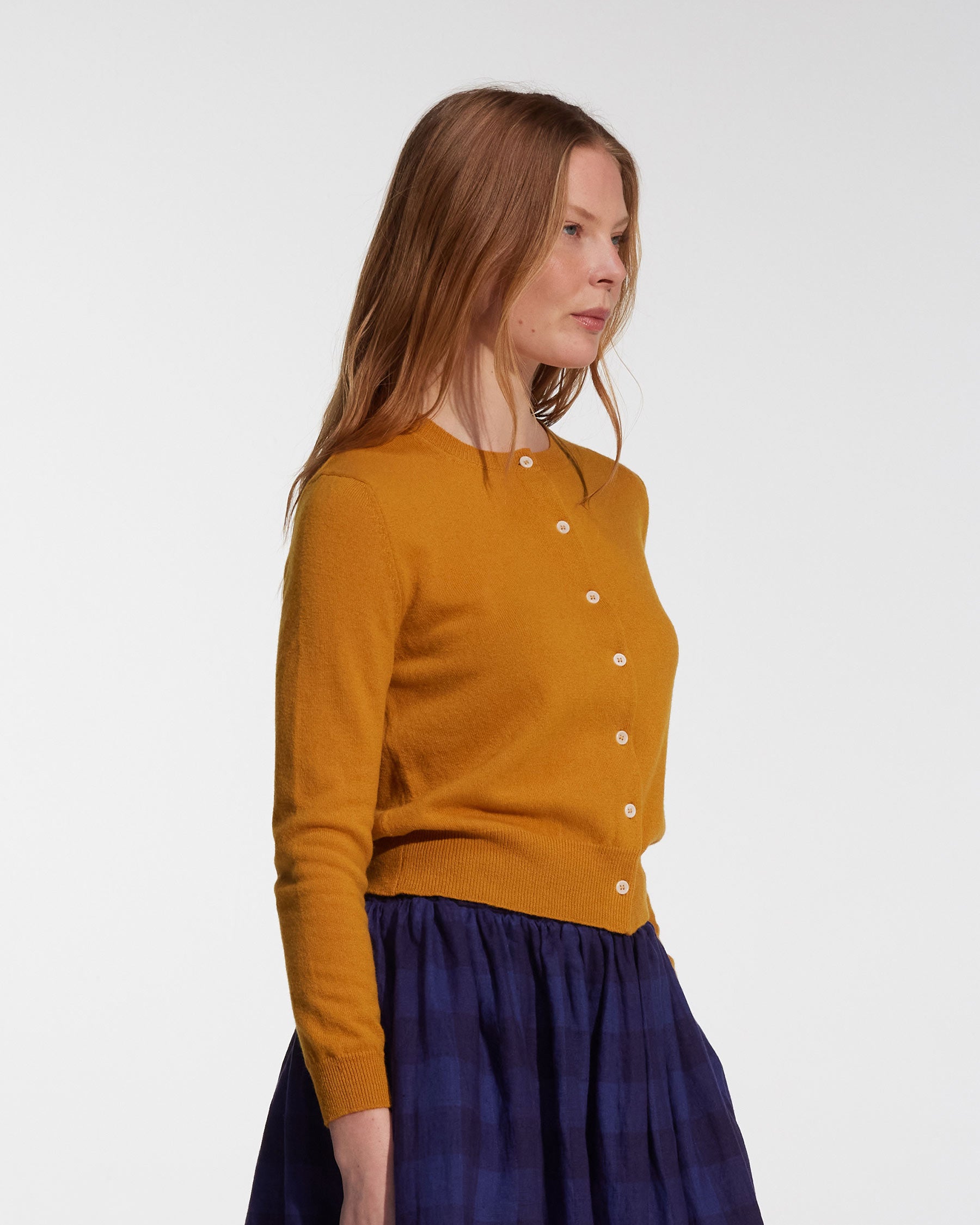 A woman with long light brown hair wears the Pyne and Smith Clothiers Merino Cardigan in Coleman's Yellow with a blue plaid skirt, standing against a plain white background.