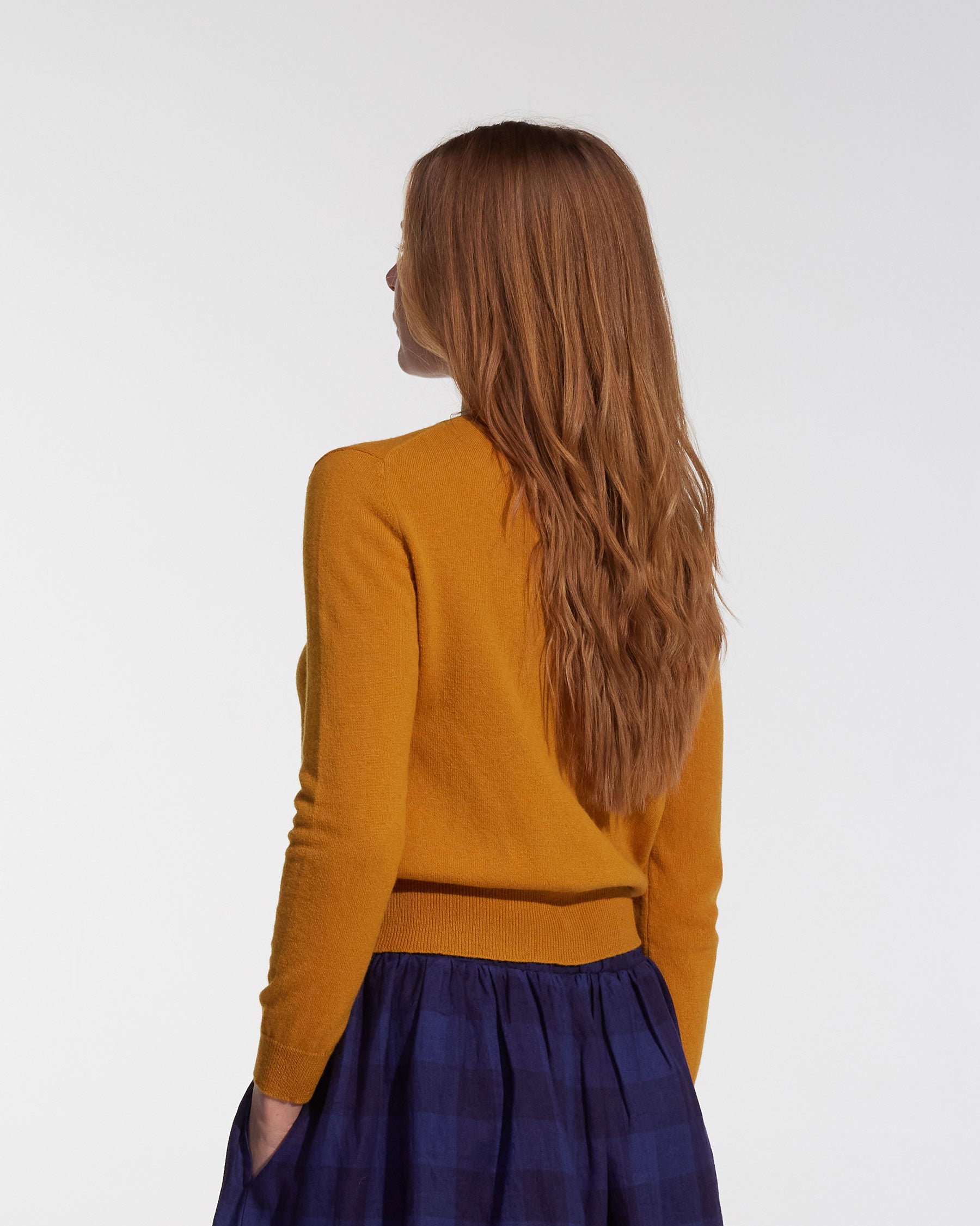 A woman with long, light brown hair faces away, wearing the Pyne and Smith Clothiers Merino Cardigan in Coleman's Yellow and a dark blue plaid skirt against a plain white background.