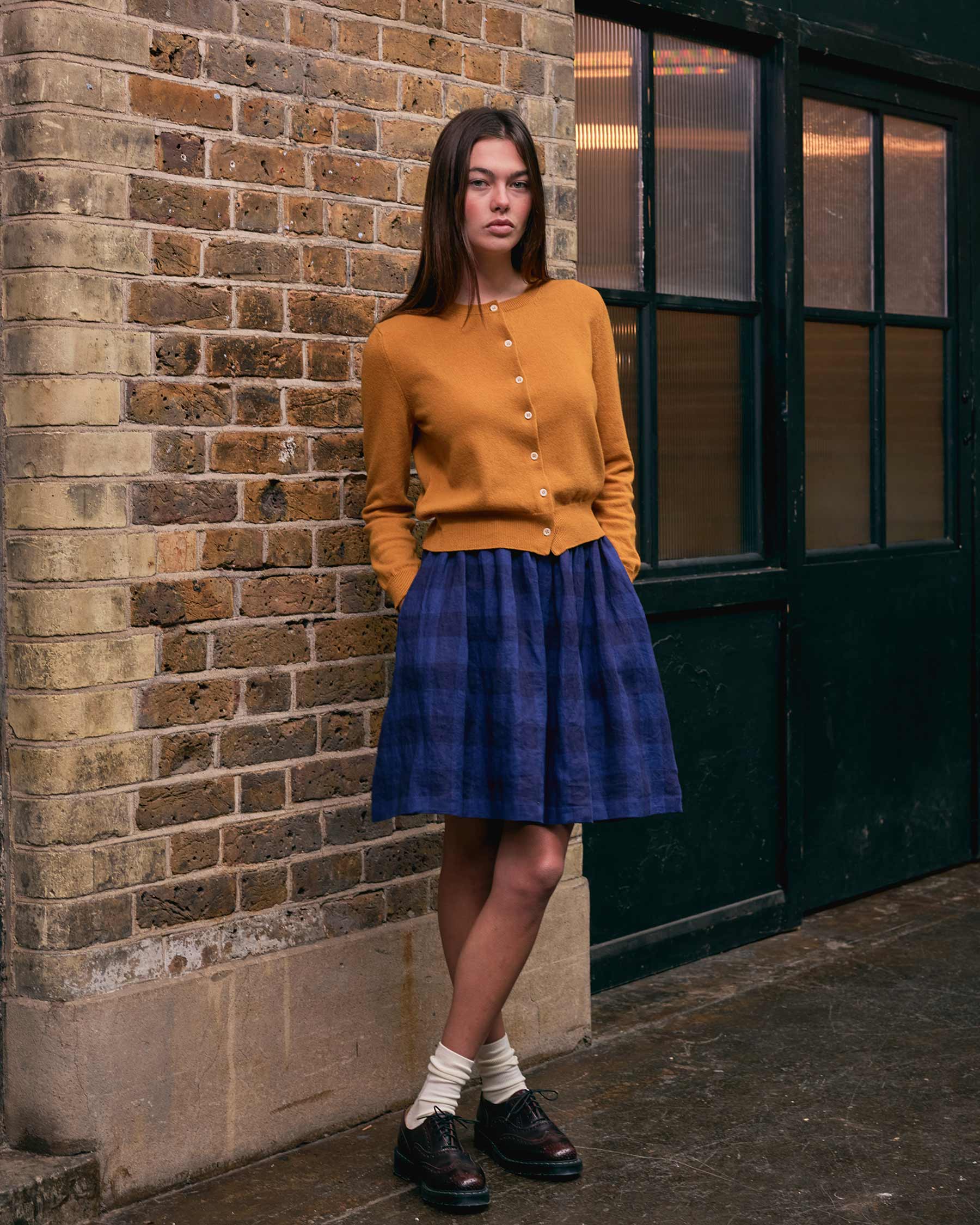 A woman stands against a brick wall wearing the Pyne and Smith Clothiers Merino Cardigan in Coleman's Yellow, paired with a blue plaid skirt, white socks, and black shoes. She looks at the camera neutrally with her hands in her pockets.