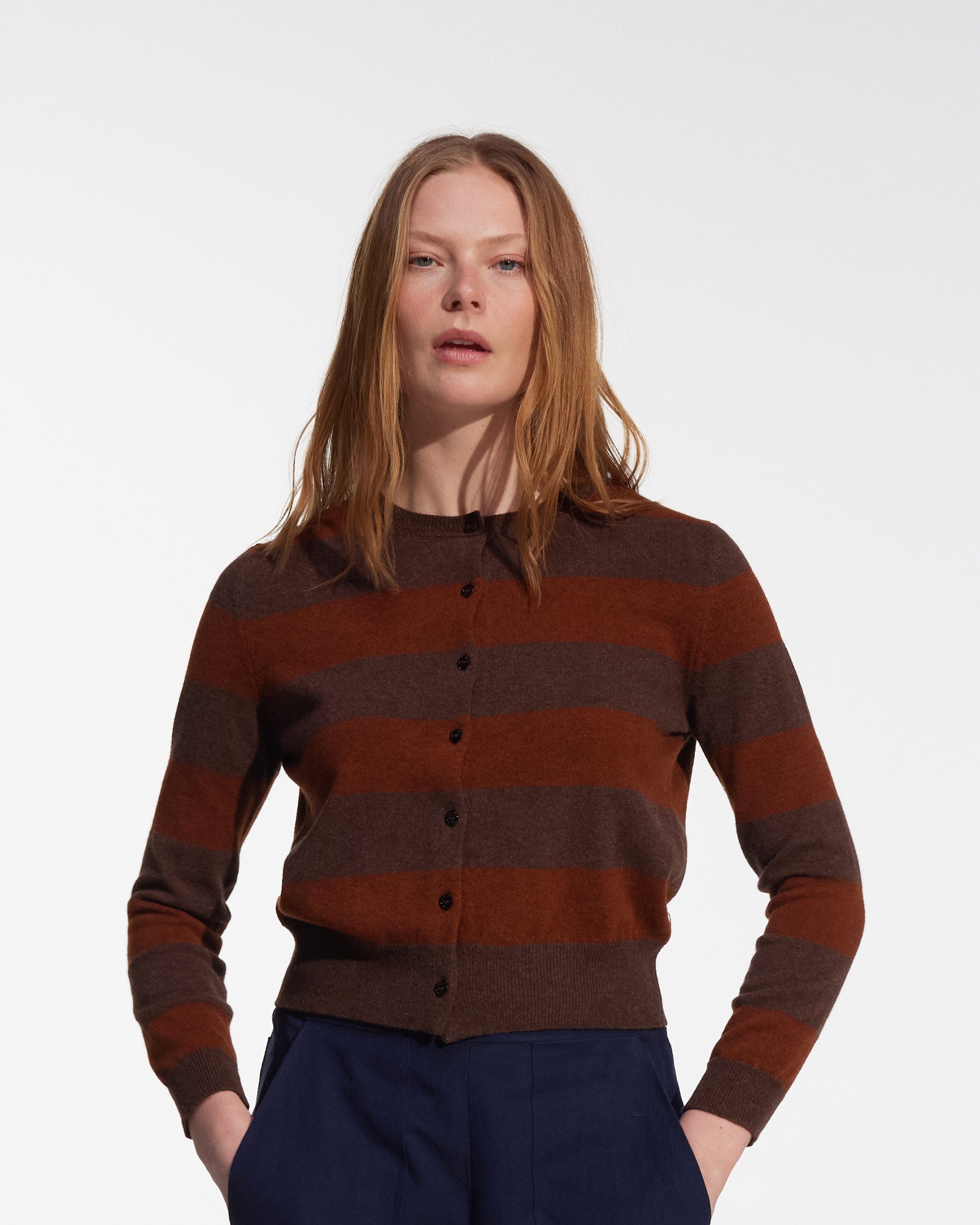 A woman with straight, light brown hair wears the Merino Cardigan in Foxtail by Pyne and Smith Clothiers— a lightweight, brown and rust striped cropped cardigan— paired with dark blue pants against a plain white background.