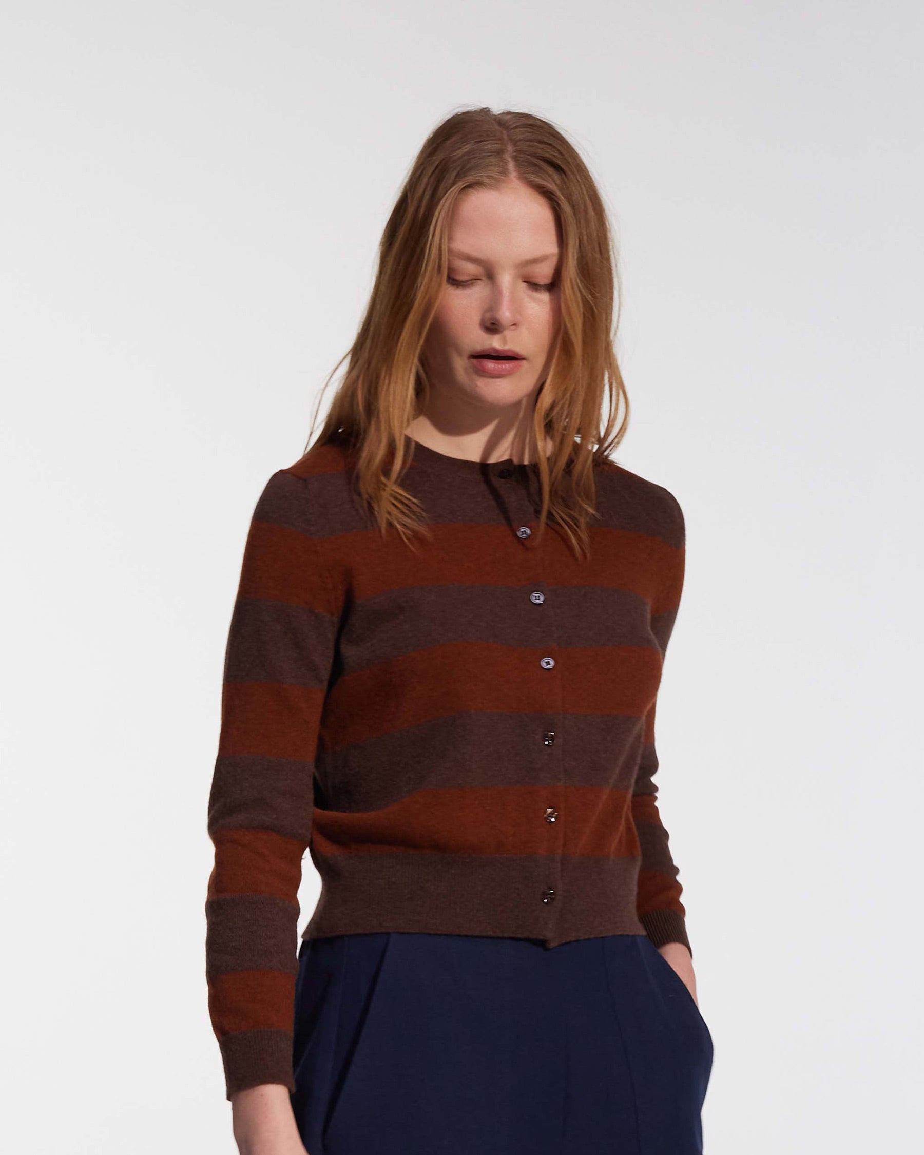 A woman with light skin and straight, light brown hair models the Pyne and Smith Clothiers Merino Cardigan in Foxtail—brown and rust striped—paired with navy blue pants, standing against a plain white background.