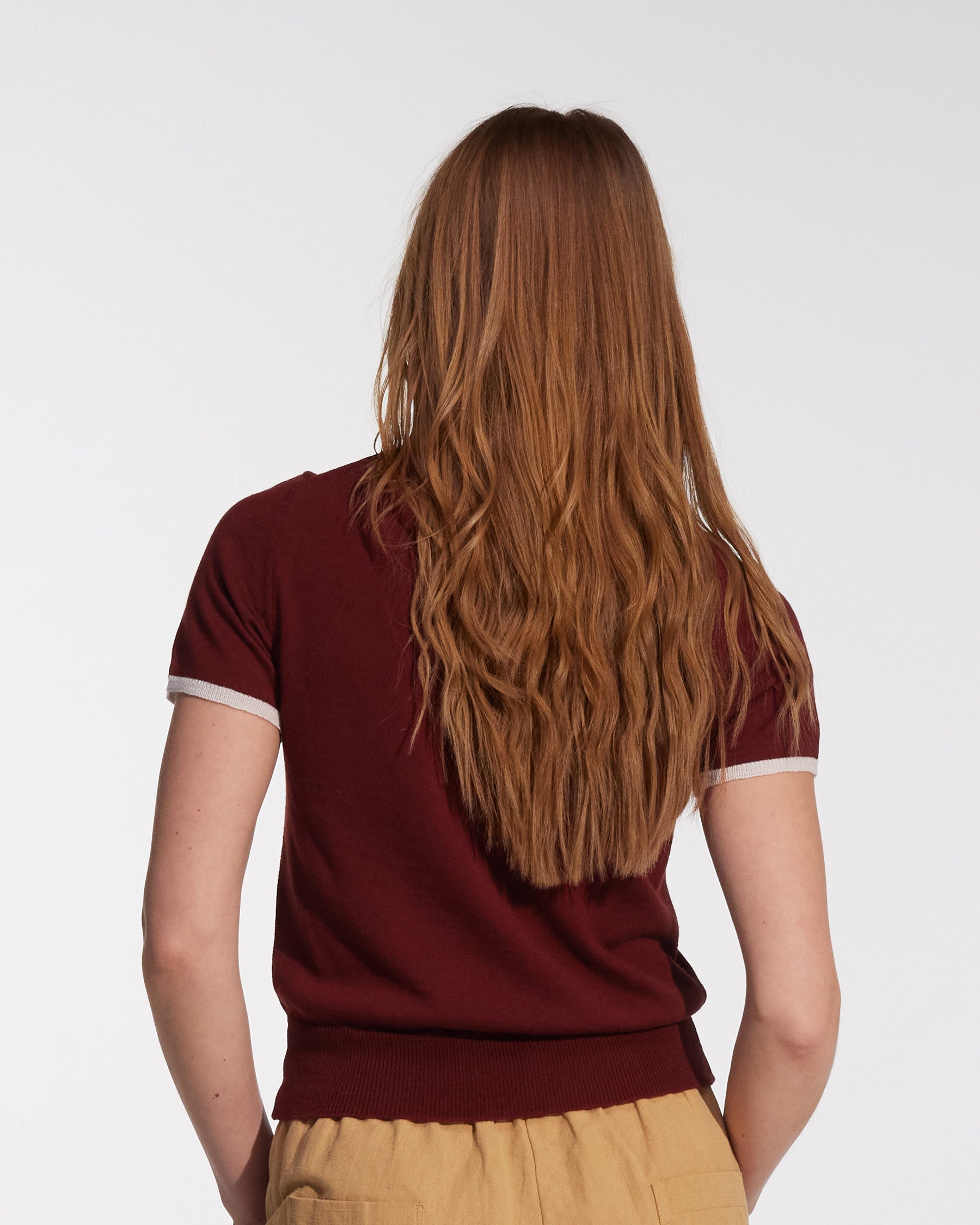 A person with long, wavy light brown hair is seen from behind, wearing the Pyne and Smith Clothiers Merino Polo Shirt in Rosso with tan pants, standing against a plain white background.