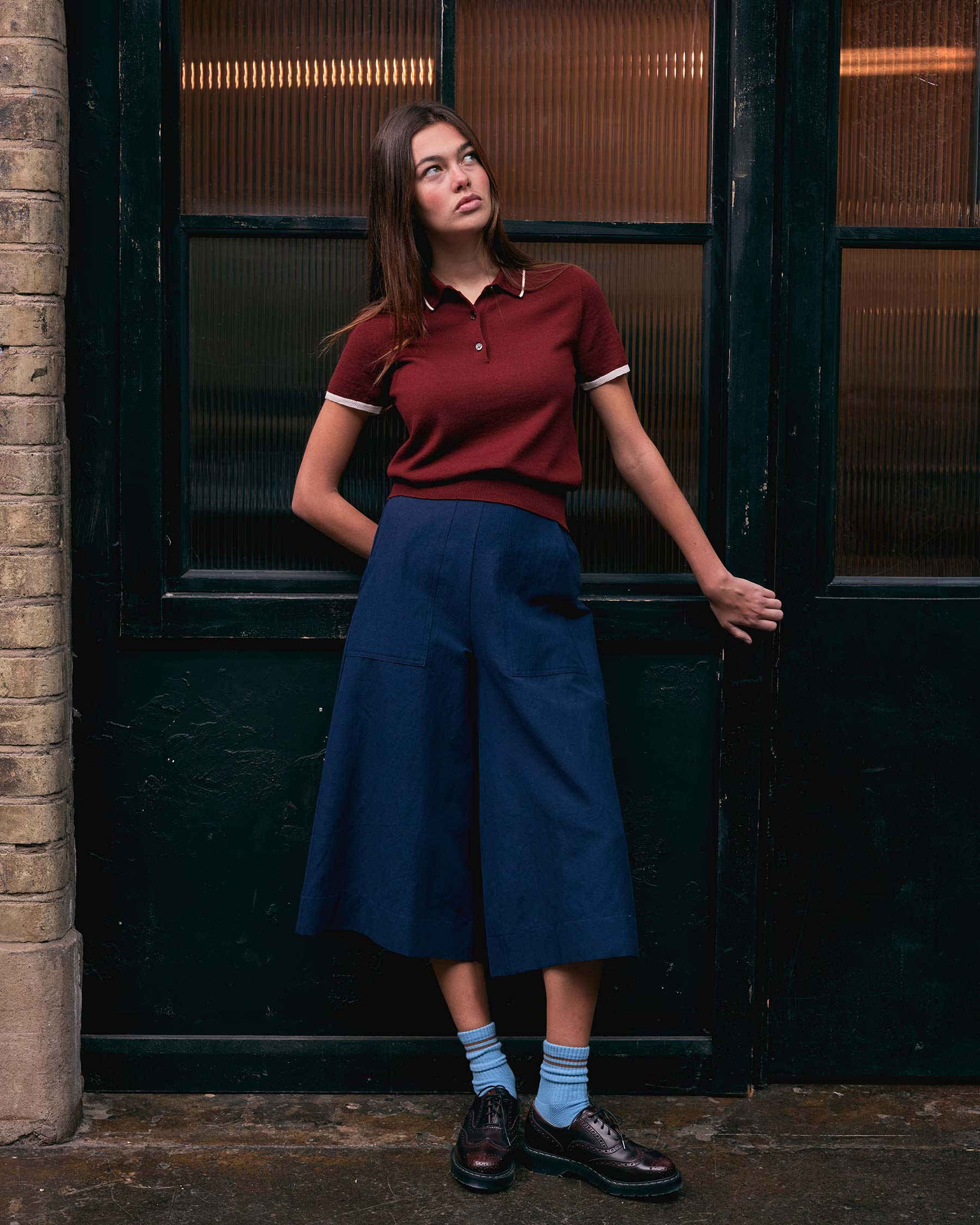 A woman with long brown hair stands before dark double doors, gazing upward with one hand in her pocket. She wears a slim-fitting Merino Polo Shirt in Rosso by Pyne and Smith Clothiers, a navy ankle-length skirt, blue socks, and dark shoes.
