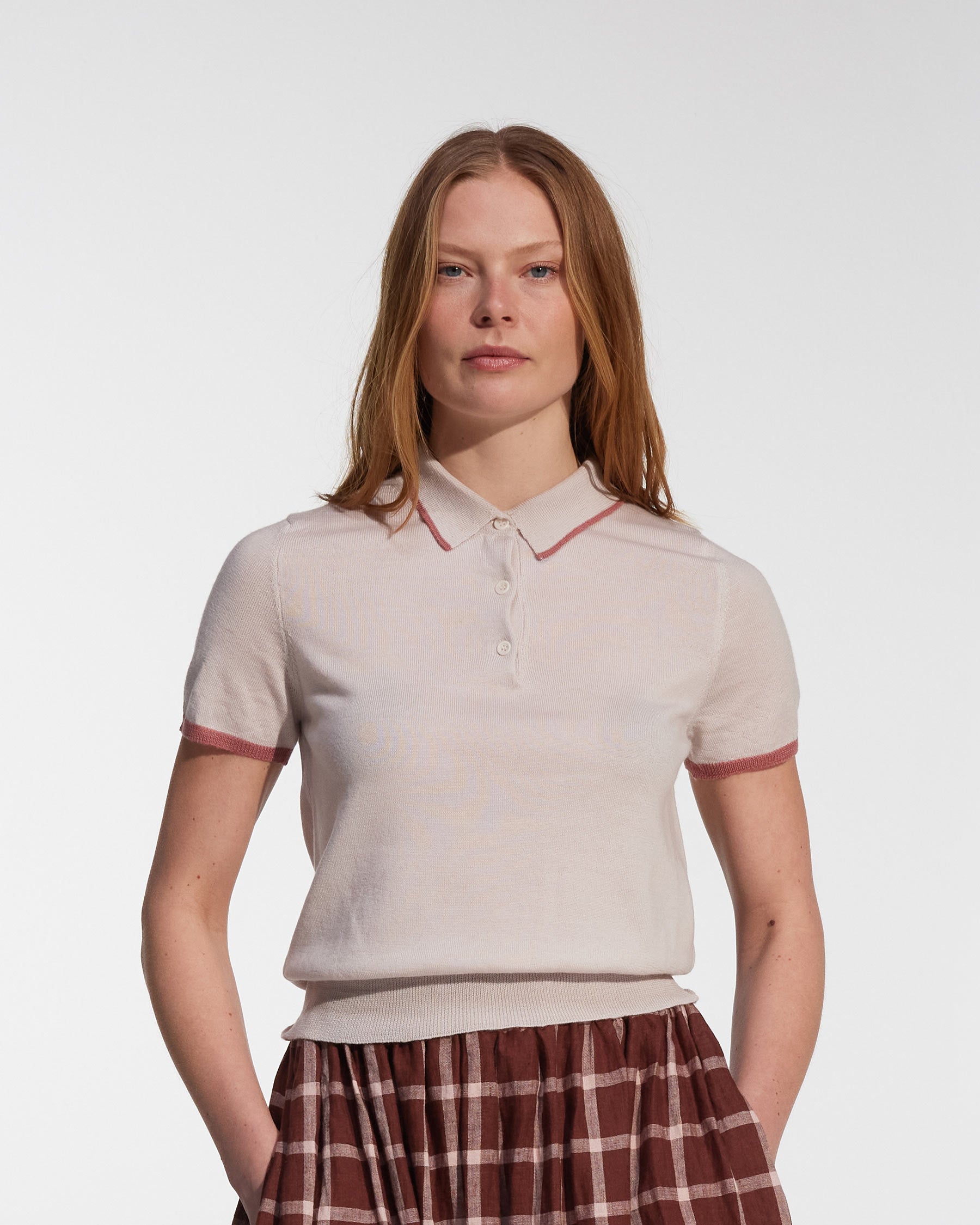A woman with long red hair wears the Pyne and Smith Clothiers Merino Polo Shirt in Shell Pink, styled slim fit, paired with a maroon plaid skirt, standing against a plain white background.