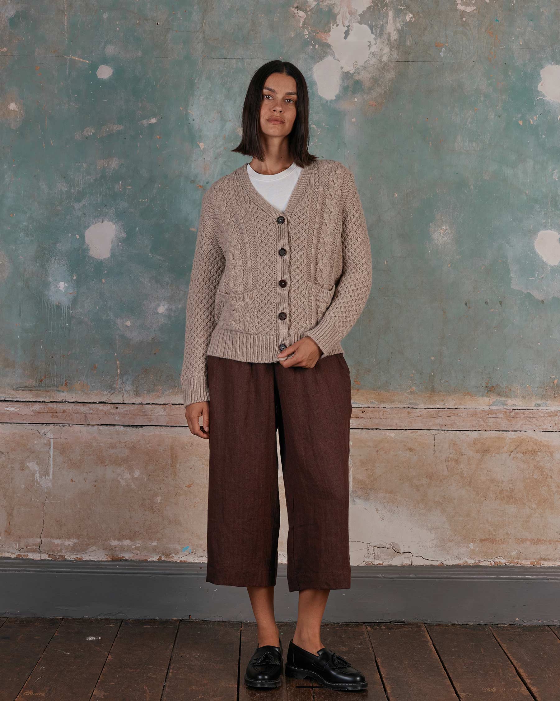A woman stands indoors by a weathered wall, wearing the Pyne and Smith Clothiers Merino Wool Cable Knit Cardigan with Pockets in Wheaten over a white shirt, brown wide-legged pants, and black shoes on a wooden floor.