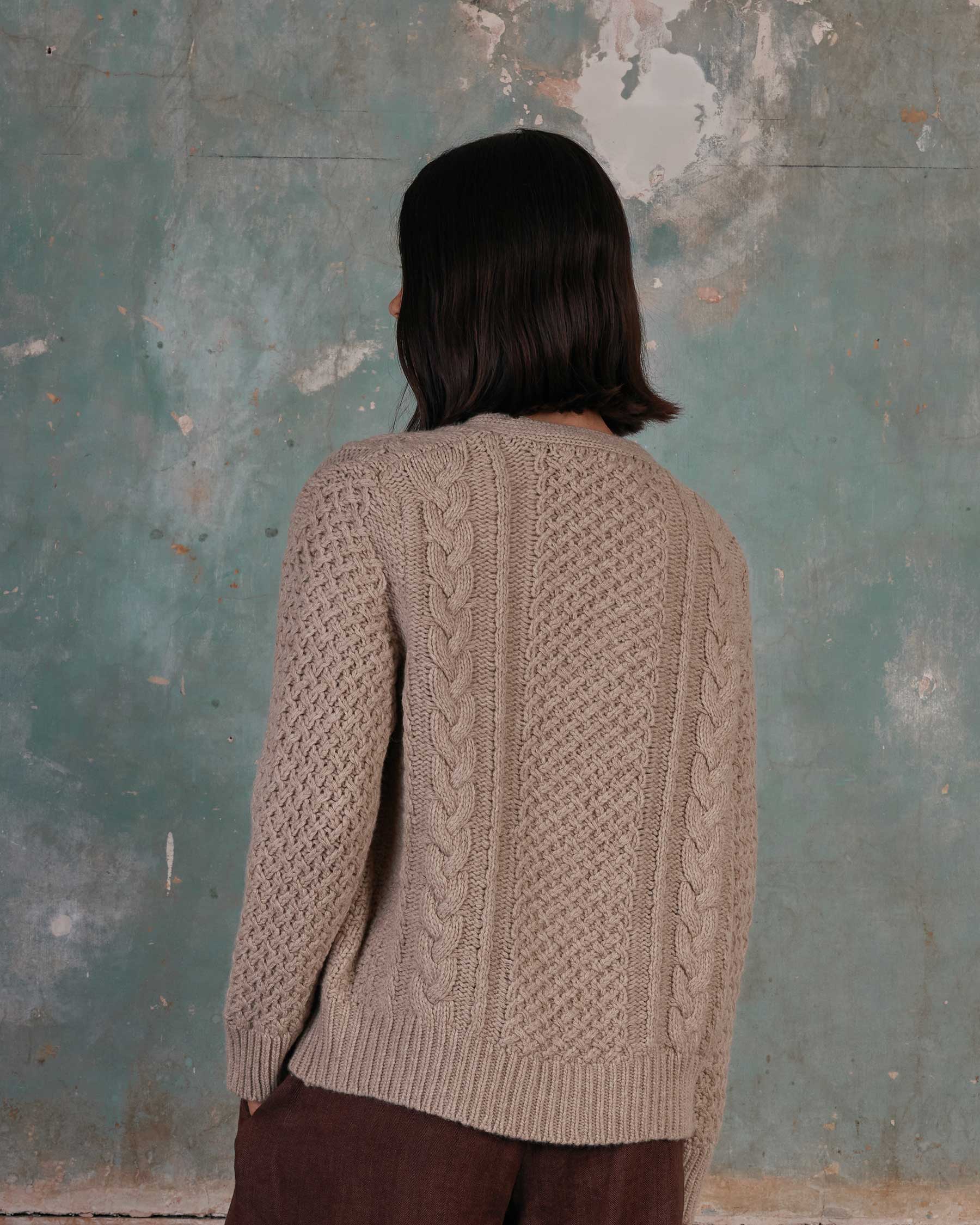 A person with shoulder-length dark hair, wearing the Pyne and Smith Clothiers Merino Wool Cable Knit Cardigan with Pockets in Wheaten, stands with their back to the camera against a distressed teal and white wall.