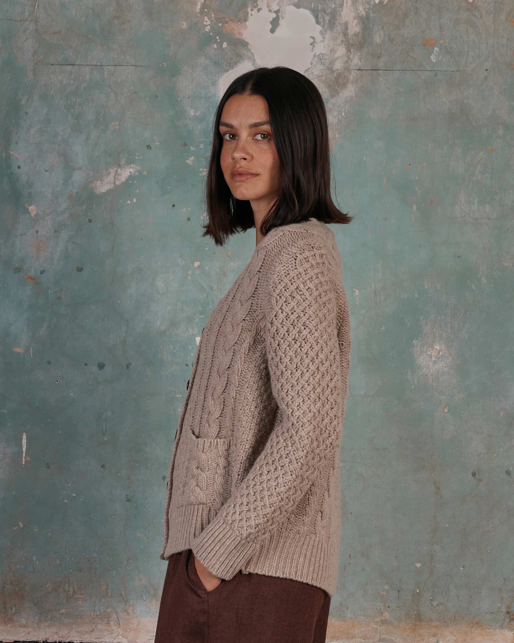A woman with straight, dark hair and a neutral expression stands sideways against a weathered blue-green wall, wearing Pyne and Smith Clothiers Merino Wool Cable Knit Cardigan with Pockets in Wheaten and brown pants.