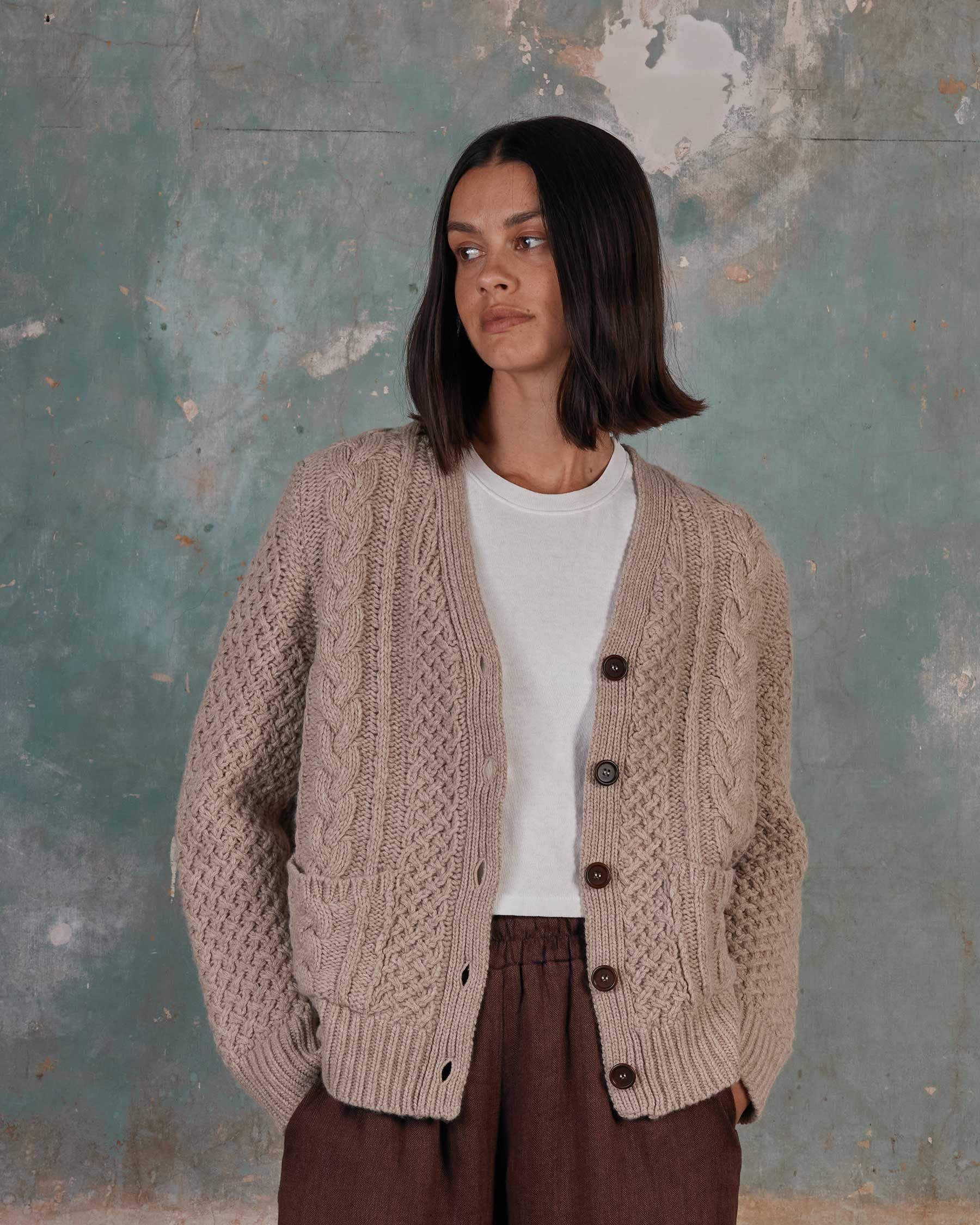 A woman with straight shoulder-length dark hair wears the Pyne and Smith Clothiers Merino Wool Cable Knit Cardigan with Pockets in Wheaten over a white tee and brown pants, standing against a textured blue-green wall and looking to the side.